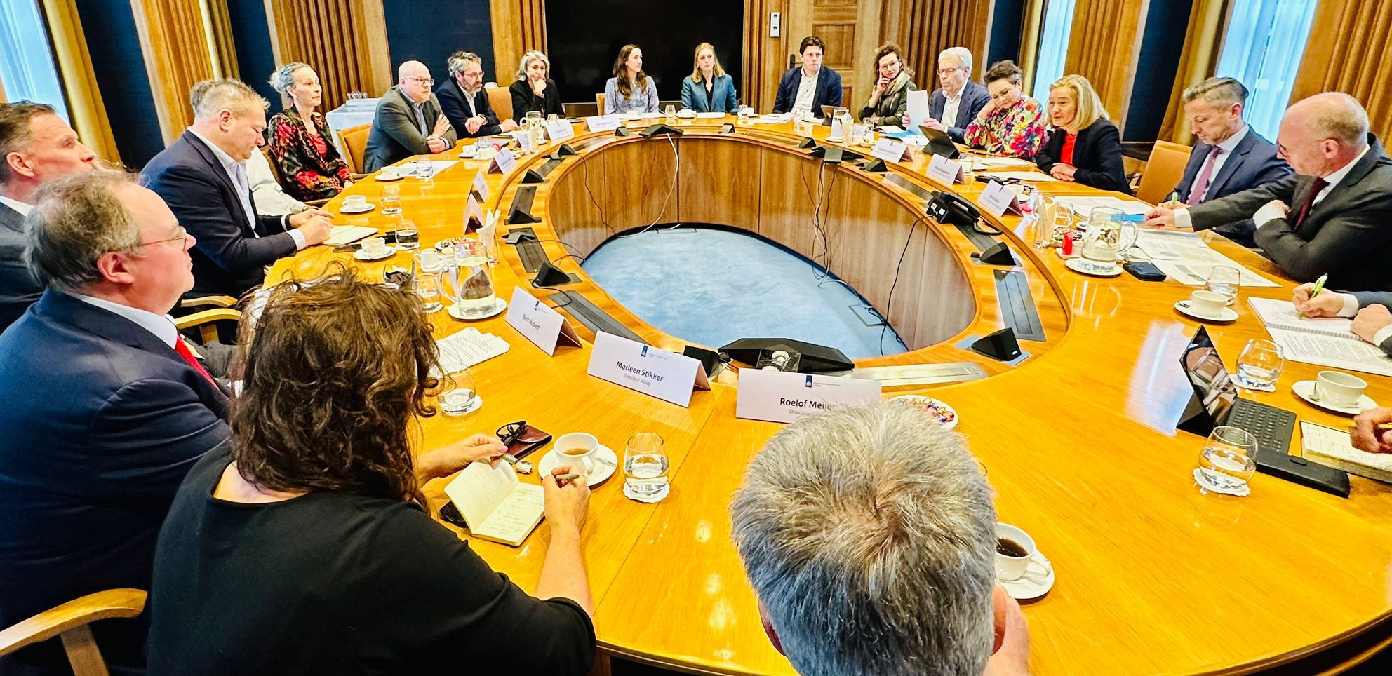 Ovale tafel vol mensen met naambordjes, met aan de rechterkant de minister en staatssecretaris